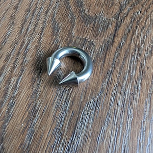 May include: A circular silver-toned metal horseshoe barbell body jewelry piece featuring two pointed cone-shaped ends. The smooth, polished surface of the circular ring reflects light, and the two spikes are positioned facing each other at the opening.