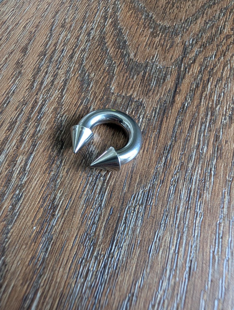 May include: A circular silver-toned metal horseshoe barbell body jewelry piece featuring two pointed cone-shaped ends. The smooth, polished surface of the circular ring reflects light, and the two spikes are positioned facing each other at the opening.