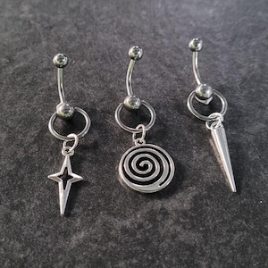 Puede incluir: Tres anillos de ombligo plateados con diferentes colgantes. Los colgantes son una estrella, una espiral y una punta.