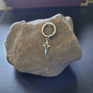 May include: A silver-colored earring with a star-shaped charm. The earring is a hoop style with a small star pendant hanging from it. The star has a four-pointed design. The earring is displayed on a gray rock.