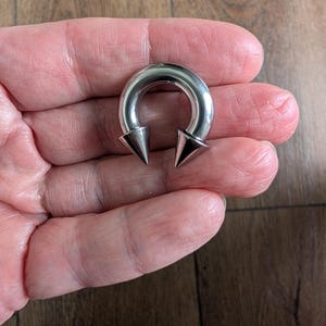 May include: A circular horseshoe-shaped body jewelry piece made of polished silver-toned metal. The item features two pointed cone-shaped ends that screw onto the curved bar, creating a symmetrical design with a smooth, reflective metallic finish.