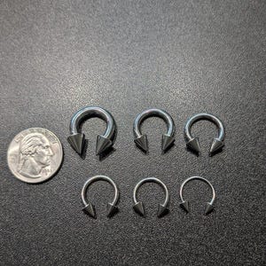 May include: A collection of five silver-toned circular barbell body jewelry pieces with pointed cone ends, arranged in two rows next to a United States quarter coin for scale, showing varying sizes of the circular hoops and the attached spikes.