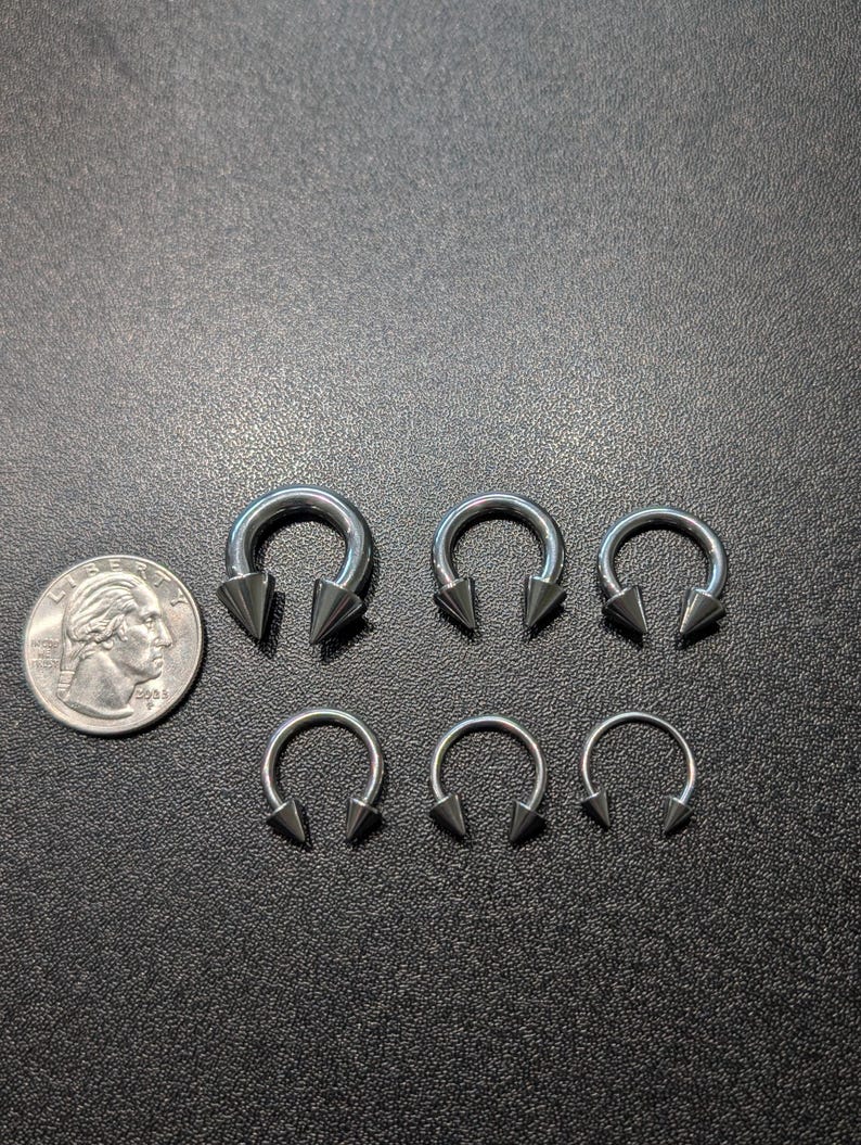 May include: A collection of five silver-toned circular barbell body jewelry pieces with pointed cone ends, arranged in two rows next to a United States quarter coin for scale, showing varying sizes of the circular hoops and the attached spikes.