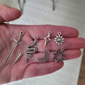 May include: A collection of silver-colored charms, including a sword, a dagger, a snake, a star, a sun, a crescent moon, and a decorative sunburst. These charms are likely for jewelry making or other craft projects.