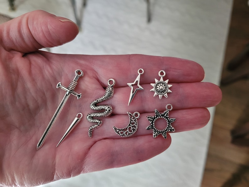 May include: A collection of silver-colored charms, including a sword, a dagger, a snake, a star, a sun, a crescent moon, and a decorative sunburst. These charms are likely for jewelry making or other craft projects.