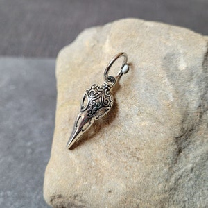 May include: A silver skull pendant with intricate details, featuring a loop for attaching a chain. The pendant is on a gray textured surface.