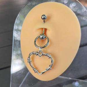 May include: A silver barbell with a silver ring and a barbed wire heart charm. The barbell is inserted into a fake earlobe.