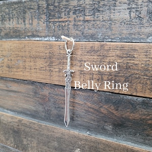 May include: Silver sword belly ring with intricate details. The ring is hanging from a wooden surface.
