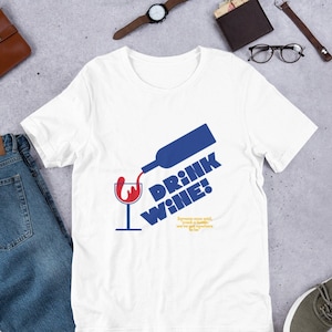 Op de afbeelding: Wit T-shirt met een blauwe en rode afbeelding van een wijnfles die in een glas wordt gegoten. De tekst "DRINK WINE!" is in vetgedrukte, blauwe letters geschreven. De quote "Someone once said, 'crack a bottle - we've got nowhere to be'" is in kleinere, gele letters geschreven.