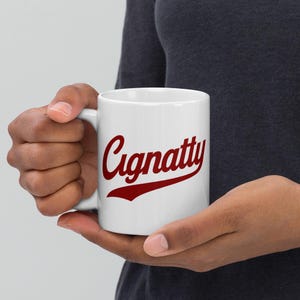 Könnte beinhalten: Weißer Keramikbecher mit dem Wort "Cignatty" in roter Schrift. Der Becher wird von einer Person mit dunkler Haut gehalten. Der Becher hat einen Griff auf der linken Seite.