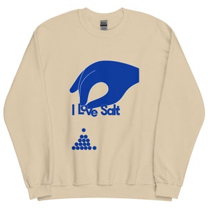 Op de afbeelding: Een beige sweatshirt met een blauwe hand die een zoutvaatje vasthoudt en de tekst "I Love Salt". Een stapel blauwe zoutkristallen bevindt zich onder de tekst.