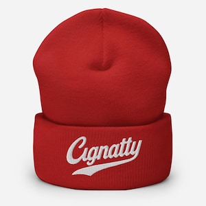 Könnte beinhalten: Rote Beanie-Mütze mit dem Wort "Cignatty" in weißer Schrift auf der Vorderseite gestickt. Die Mütze besteht aus weichem Strickmaterial und hat einen umgeschlagenen Rand.