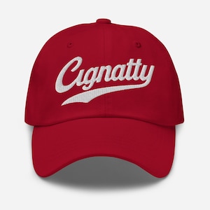 Könnte beinhalten: Rote Baseballkappe mit dem weißen, gestickten Wort "Cignatty" in Schreibschrift auf der Vorderseite. Die Kappe hat einen gebogenen Schirm und einen Stoffriemenverschluss.