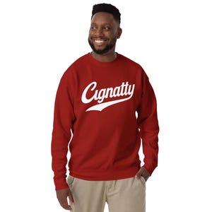 Könnte beinhalten: Ein rotes Sweatshirt mit dem Wort "Cignatty" in weißer Schrift auf der Brust. Das Sweatshirt ist einfarbig und hat einen Rundhalsausschnitt. Die Person auf dem Bild trägt eine khakifarbene Hose.