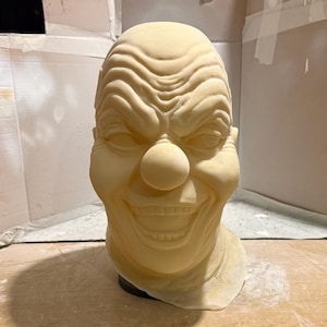 May include: A pale yellow clown mask with a large red nose and a wide, unsettling grin. The mask has detailed wrinkles and is designed to be worn. The overall theme is horror and Halloween.