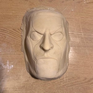 May include: A light beige mask with a stern expression. The mask has defined facial features, including a prominent nose, furrowed brow, and a defined jawline. The mask is likely for costume or artistic purposes.