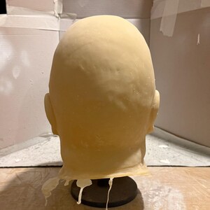 Peut inclure: Un masque de t&ecirc;te chauve jaune p&acirc;le avec des oreilles visibles, mont&eacute; sur un support noir. Le masque a une texture lisse et est probablement fait d'un mat&eacute;riau souple. L'arri&egrave;re de la t&ecirc;te est visible, et le masque semble &ecirc;tre un accessoire ou un &eacute;l&eacute;ment de costume.