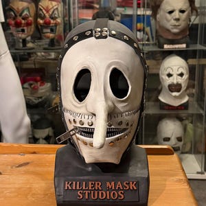 May include: A white mask with a long nose and a zipper mouth, secured with a black leather strap and metal studs. The mask is mounted on a black base with "KILLER MASK STUDIOS" in brown lettering. Other masks are visible in the background.