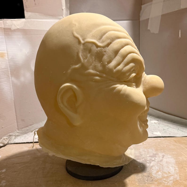 Peut inclure: Masque sculpt&eacute; jaune p&acirc;le repr&eacute;sentant un visage grotesque, avec un grand nez, un front rid&eacute; et un l&eacute;ger sourire. Le masque est mont&eacute; sur une base noire, sugg&eacute;rant qu'il s'agit d'un accessoire ou d'un objet d&eacute;coratif. Les d&eacute;tails sont finement rendus, lui donnant un aspect r&eacute;aliste.