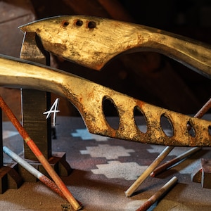 May include: Two rustic, metal, knife-like objects with handles, resting on wooden blocks. The objects are painted in a gold and brown color with a distressed finish. The objects are labeled A and B.