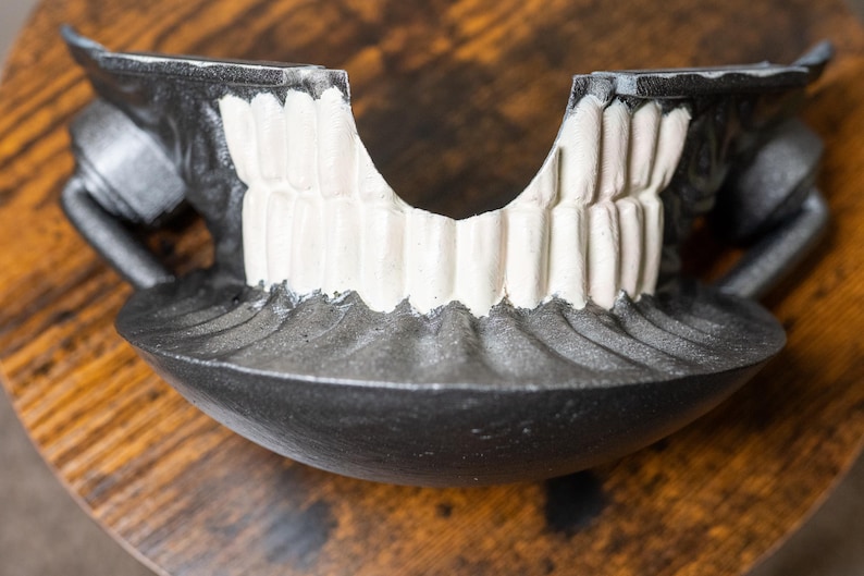 K&ouml;nnte beinhalten: Eine dunkelgraue, stilisierte Kieferknochenskulptur mit wei&szlig;en Z&auml;hnen. Die Skulptur hat eine schalenf&ouml;rmige Basis und zwei seitliche Griffe. Die Z&auml;hne sind detailliert und das Gesamtdesign hat ein gotisches oder Halloween-Thema.