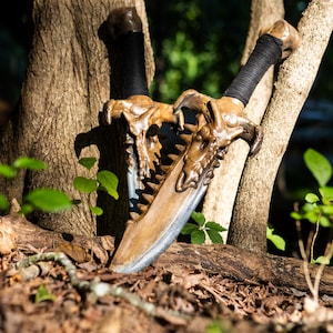 May include: Two decorative swords with skull-shaped handles and a silver blade. The swords are made of wood and have a brown and black finish. The swords are lying on the ground in a forest setting.