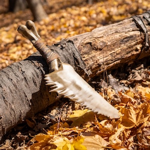 May include: A bone-like dagger with a leather wrap handle lies on a fallen log in a forest setting. The dagger has a serrated edge and a pointed tip. The log is surrounded by fallen leaves in shades of brown and yellow.