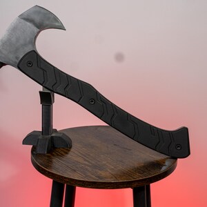 May include: A black and silver axe with a curved blade and a textured handle rests on a small wooden stool. The axe has a silver blade and a black handle with a geometric pattern. The stool has a dark wooden seat and black legs.