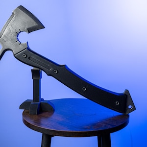 May include: A black tactical tomahawk axe with a curved handle and a hammerhead design. The axe head features a circular cutout and is mounted on a small black stand. The background is a gradient of blue.