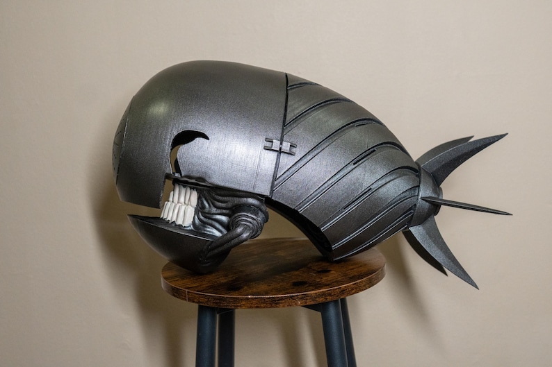 K&ouml;nnte beinhalten: Ein metallisch grauer Helm mit einem sch&auml;delartigen Gesicht und einer flossenartigen Struktur am R&uuml;cken. Der Helm liegt auf einem kleinen, runden Holzschemel. Das Design weist einen Klappabschnitt und detaillierte Texturen auf.