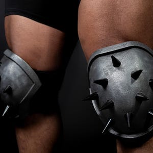Könnte beinhalten: Paar graue Kniepolster mit Stacheln. Die Polster bestehen aus einem metallischen Material und weisen mehrere schwarze Stacheln auf. Das Bild zeigt die Polster an den Knien einer Person, mit dunklem Hintergrund.