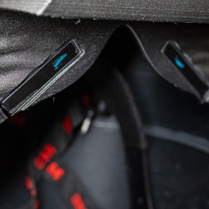 K&ouml;nnte beinhalten: Nahaufnahme eines schwarzen, strukturierten Objekts mit geschwungenem Design. Zwei kleine, rechteckige schwarze Akzente mit blauem Design sind sichtbar. Rote und schwarze Riemen sind teilweise sichtbar.