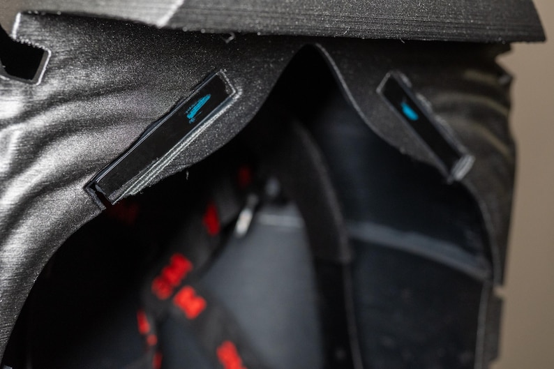 K&ouml;nnte beinhalten: Nahaufnahme eines schwarzen, strukturierten Objekts mit geschwungenem Design. Zwei kleine, rechteckige schwarze Akzente mit blauem Design sind sichtbar. Rote und schwarze Riemen sind teilweise sichtbar.
