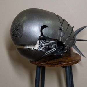 K&ouml;nnte beinhalten: Eine metallisch graue Skulptur einer Kreatur mit rundem Kopf, offenem Mund mit wei&szlig;en Z&auml;hnen und einem stacheligen Schwanz. Die Skulptur hat eine strukturierte Oberfl&auml;che und steht auf einem kleinen Holzschemel.
