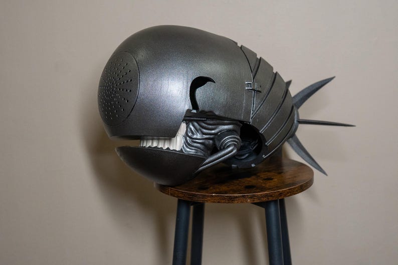 K&ouml;nnte beinhalten: Eine metallisch graue Skulptur einer Kreatur mit rundem Kopf, offenem Mund mit wei&szlig;en Z&auml;hnen und einem stacheligen Schwanz. Die Skulptur hat eine strukturierte Oberfl&auml;che und steht auf einem kleinen Holzschemel.