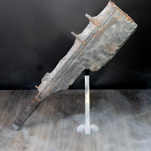 May include: A large, heavy-duty metal cleaver with a weathered, textured grey finish and visible rust spots. The blade features a thick spine with metal brackets, and the handle is wrapped in dark, worn material, mounted on a vertical stand.