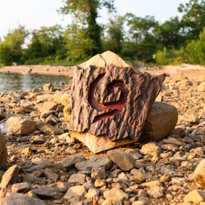May include: A pentagonal stone plaque featuring a textured, bark-like brown surface with a carved, recessed spiral symbol in the center. The carving reveals a smooth, deep red interior surface within the spiral design, set against a rocky shoreline.