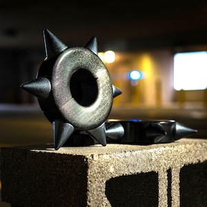 Könnte beinhalten: Zwei schwarze, mit Spikes besetzte Metallringe mit einem Loch in der Mitte, die auf einem grauen Betonblock liegen.