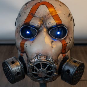 May include: A full-face mask featuring a weathered off-white finish with prominent orange stripes and dark distressed detailing. The mask includes two circular side filters and a central vent, with bright blue LED lights glowing from the eye sockets.