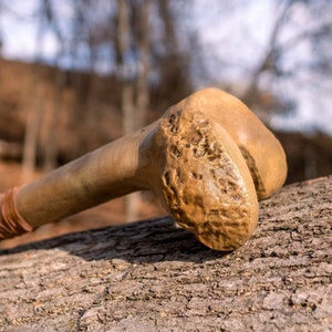 Op de afbeelding: Een houten knuppel met een getextureerd oppervlak en een bruine leren wikkeling. De knuppel rust op een stuk boomschors.