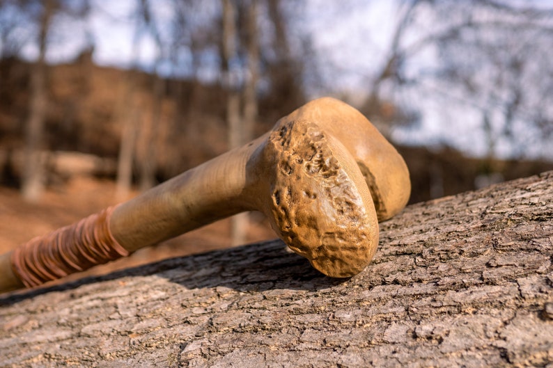 Op de afbeelding: Een houten knuppel met een getextureerd oppervlak en een bruine leren wikkeling. De knuppel rust op een stuk boomschors.