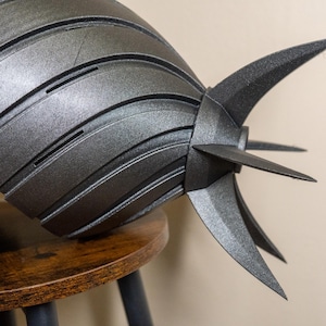 K&ouml;nnte beinhalten: Ein metallisch graues, segmentiertes Objekt mit spitzen Flossen, m&ouml;glicherweise ein Helm oder ein dekoratives St&uuml;ck. Das Objekt liegt auf einem kleinen, runden Holzschemel. Das Design weist geschwungene Linien und scharfe Winkel auf und erzeugt eine futuristische &Auml;sthetik.