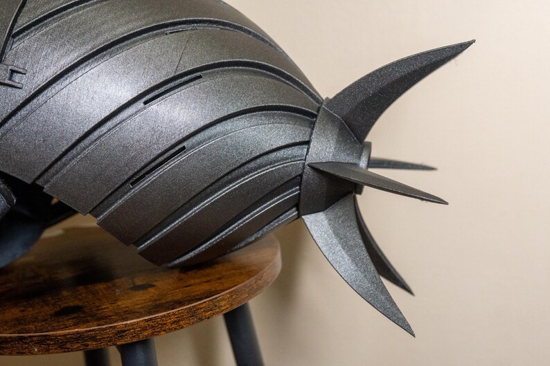 K&ouml;nnte beinhalten: Ein metallisch graues, segmentiertes Objekt mit spitzen Flossen, m&ouml;glicherweise ein Helm oder ein dekoratives St&uuml;ck. Das Objekt liegt auf einem kleinen, runden Holzschemel. Das Design weist geschwungene Linien und scharfe Winkel auf und erzeugt eine futuristische &Auml;sthetik.