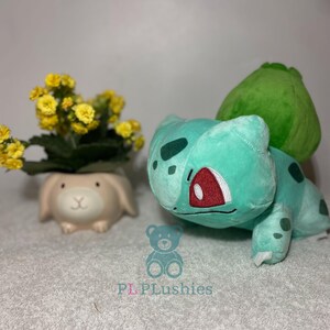 Könnte beinhalten: Ein grünes und blaues Plüschtier des Pokémon-Charakters Bulbasaur. Das Spielzeug hat ein großes grünes Blatt auf dem Rücken und rote Augen. Das Spielzeug sitzt auf einer weißen Oberfläche mit einer kleinen Topfpflanze im Hintergrund.