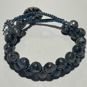 Puede incluir: Un brazalete de cuentas azul y plateado con un dije de flor plateada. Las cuentas son de color gris oscuro con destellos iridiscentes.
