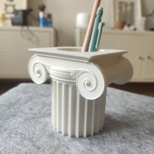 Pode incluir: Porta-lápis de cerâmica branca em forma de coluna grega em miniatura com um eixo canelado e volutas decorativas.