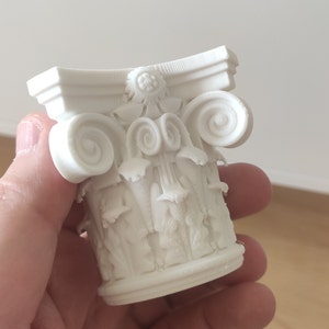 Op de afbeelding: Witte keramische miniatuur kolomkapiteel met ingewikkelde krulwerk en bloemdetails. De kapiteel is een decoratief element voor huisdecoratie of modelbouw.