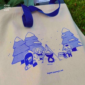 May include: Cream canvas tote bag with blue handles and a blue illustration of a camping scene with cartoon characters and trees. The text "beyond journey's end" is printed at the bottom left. The shop's name is printed at the top left.