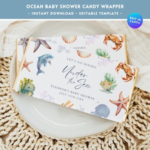 May include: A white candy bar wrapper with an ocean theme. It features a blue dolphin, a starfish, a crab, and a sea turtle. The text on the wrapper reads "Let's go diving Under the Sea" and "Eleanor's Baby Shower July 19th 20XX".
