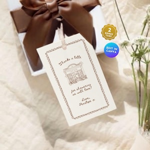 Puede incluir: Una etiqueta de regalo crema con borde marrón y texto "Thanks a latte" y "for showering us with love!". La etiqueta presenta una ilustración de café y está adjunta a una caja de regalo blanca con una cinta marrón.